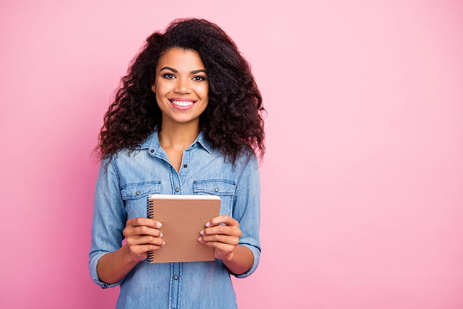 Portrait of positive cheerful african american girl holding a notebook in front of pink background